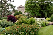 MIXED BORDERS WITH WOODEN BRIDGE AND CHURCH AT OZLEWORTH PARK, GLOUCESTERSHIRE