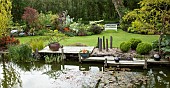 Pond with decking silver ornate balls grey slate pillars borders of colourful mature shrubs and trees in May