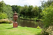 Mature trees on edge of canal large ornate terra cotta urn on plinth in June Early Summer