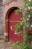RED ARCHED DOOR IN MEDIEVAL BRICK GATE WITH CLIMBING ROSES