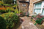 Antique chairs and a small wooden table offer seating in a quiet, sunny spot in the nulti level garden of a former farmhouse in the Yorkshire Dales.  Montbretia and sedum grow prolifically along the stone retaining walls of the garden, and various herbs and flowers grow in containers.