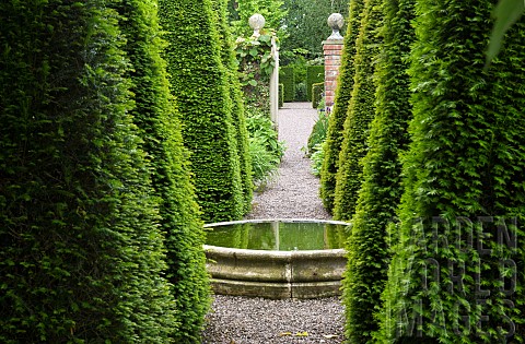 Tall_yew_spires_in_a_cruciform_carved_limestone_wellhead_in_the_Well_Garden