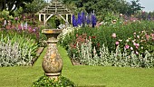 Stone sundial in lawn with twin borders of many colours and varieties of herbaceous perrenials