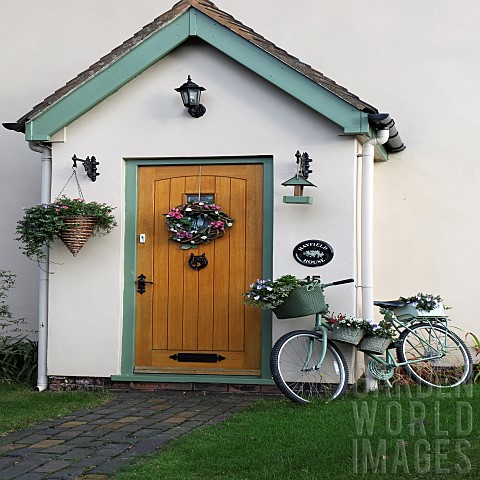 Front_garden_porch_with_wooden_door_hanging_basket_and_wreath_also_floral_displays_in_containers_on_