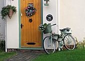 Front garden porch with wooden door hanging basket and wreath also floral displays in containers on an old pushbike