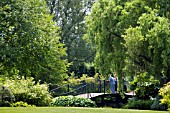 VISITORS STANDING ON THE BRIDGE OVER THE LOWER POND AT RHS GARDEN HYDE HALL,  IN JUNE.